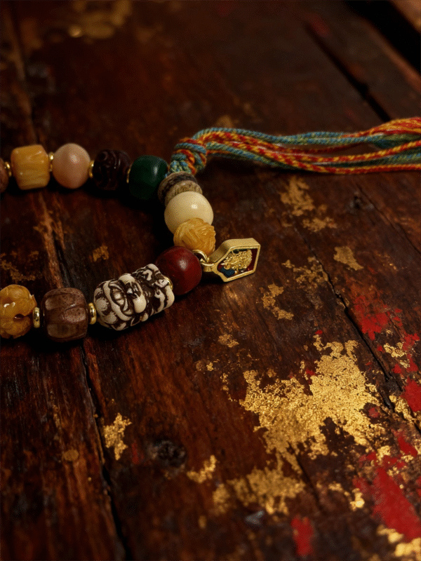 Close-up detail of the Pangu inspired After the Divide bracelet (12mm) featuring mixed beads, a colorful tassel cord and gold charm on a vintage wood background with golden texture accents