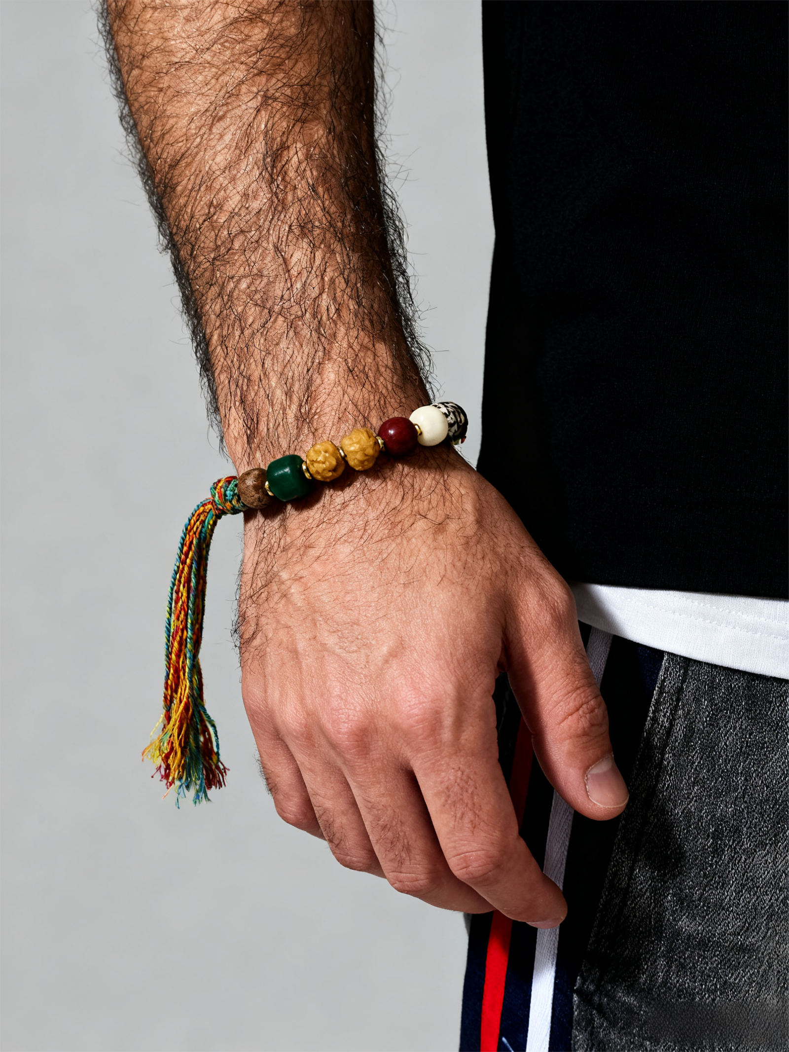 Man wearing the Pangu inspired After the Divide bracelet (12mm) featuring mixed material beads, a colorful tassel cord and gold charm on a clean background