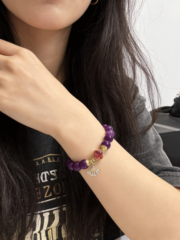 Woman wearing the Nüwa inspired Blessed Heart amethyst bracelet (11mm) with purple beads, gold accents and a red heart charm in a casual close-up wrist shot