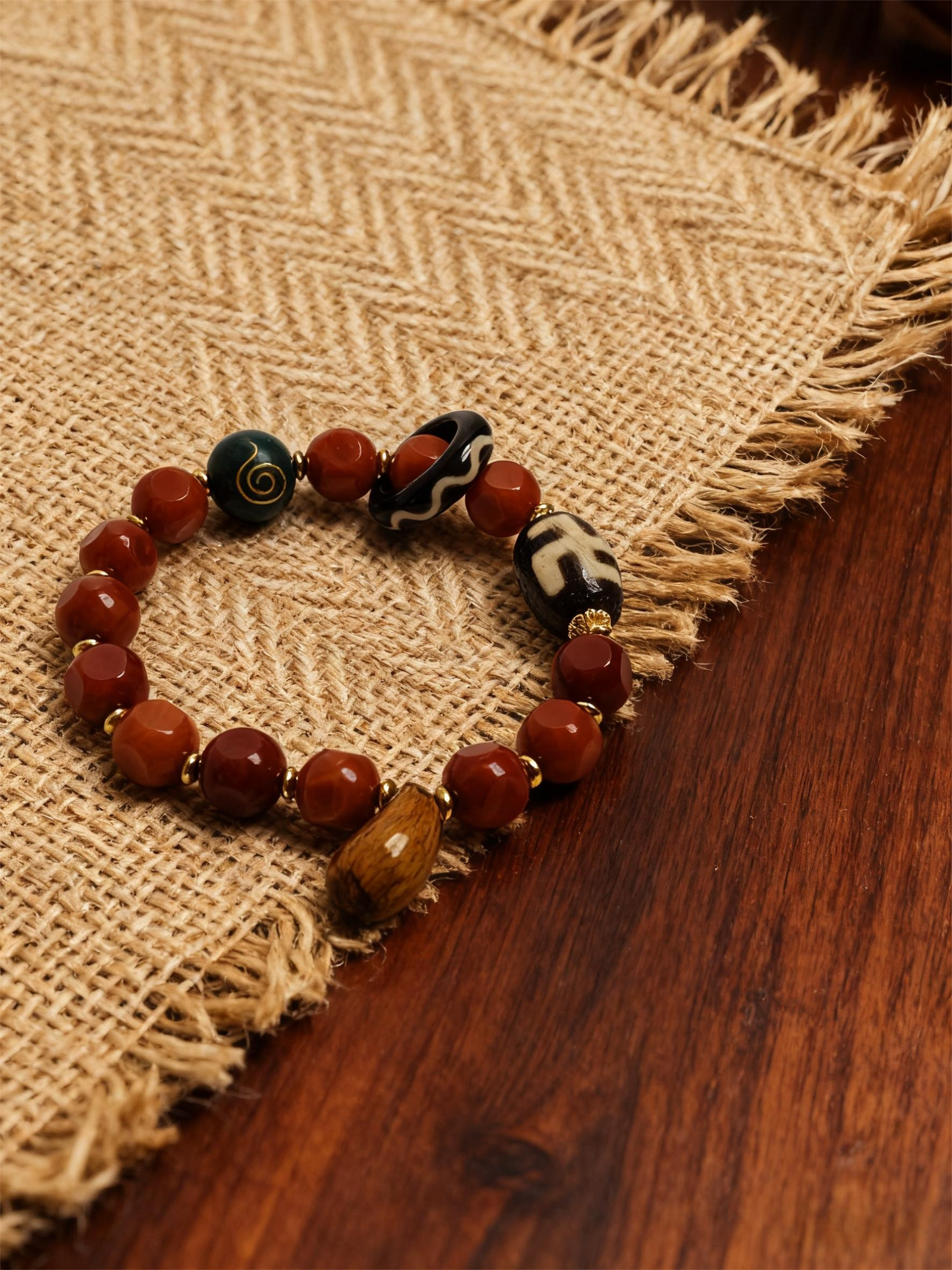 Product shot of the Xingtian inspired Bound Resolve red agate bracelet (10mm) with faceted red beads, patterned accent beads and gold details on a burlap and wooden table background