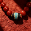 Close-up of the Bound Resolve Red Agate Bracelet featuring red agate beads and a turquoise-toned centerpiece on a textured red background.