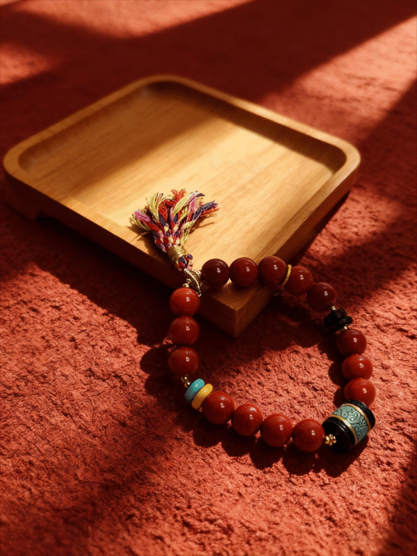 Bound Resolve Red Agate Bracelet with red agate beads and colorful tassel displayed beside a wooden tray on a textured red surface.