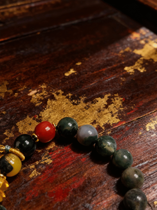Close-up of Shennong inspired apothecary bracelet featuring moss green agate beads, red accent stone, and handcrafted gold spacers on distressed wooden background