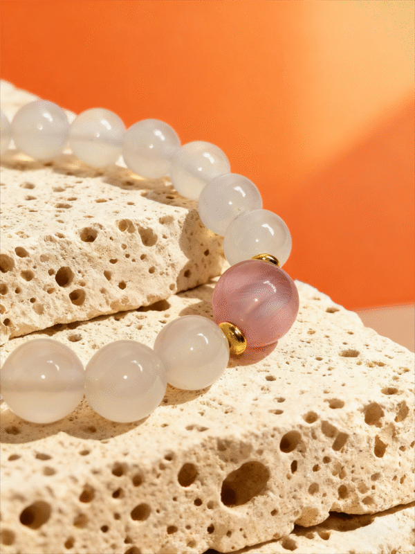 Close-up of the Chang’e inspired Goddess Moonstone & Sunrise Agate bracelet featuring translucent white moonstone beads and a soft pink sunrise agate centerpiece, accented with gold spacers, displayed on textured porous stone against a warm orange background, evoking moonlight, dawn, and feminine celestial energy.