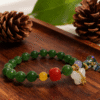 Close-up of the Crimson Herbfruit Jade Bracelet – Shennong Edition (10mm), featuring polished green jade beads, a vivid red centerpiece bead, yellow agate accents, white jade charm and gold hardware, photographed on a warm wooden surface with pine cones and botanical elements