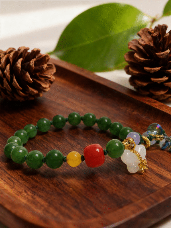 Close-up of the Crimson Herbfruit Jade Bracelet – Shennong Edition (10mm), featuring polished green jade beads, a vivid red centerpiece bead, yellow agate accents, white jade charm and gold hardware, photographed on a warm wooden surface with pine cones and botanical elements