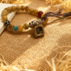 Pangu themed bone-agate bracelet laid flat with square amulet charm, natural agate beads and multicolor braided tassel on rustic woven surface