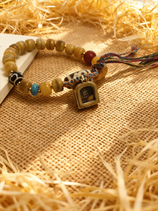 Pangu themed bone-agate bracelet laid flat with square amulet charm, natural agate beads and multicolor braided tassel on rustic woven surface
