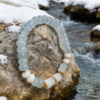 Product shot of the Jingwei inspired Quiet Oath aquamarine bracelet (6mm) displayed on a rock beside a snowy stream, featuring icy blue beads, white accents and gold details