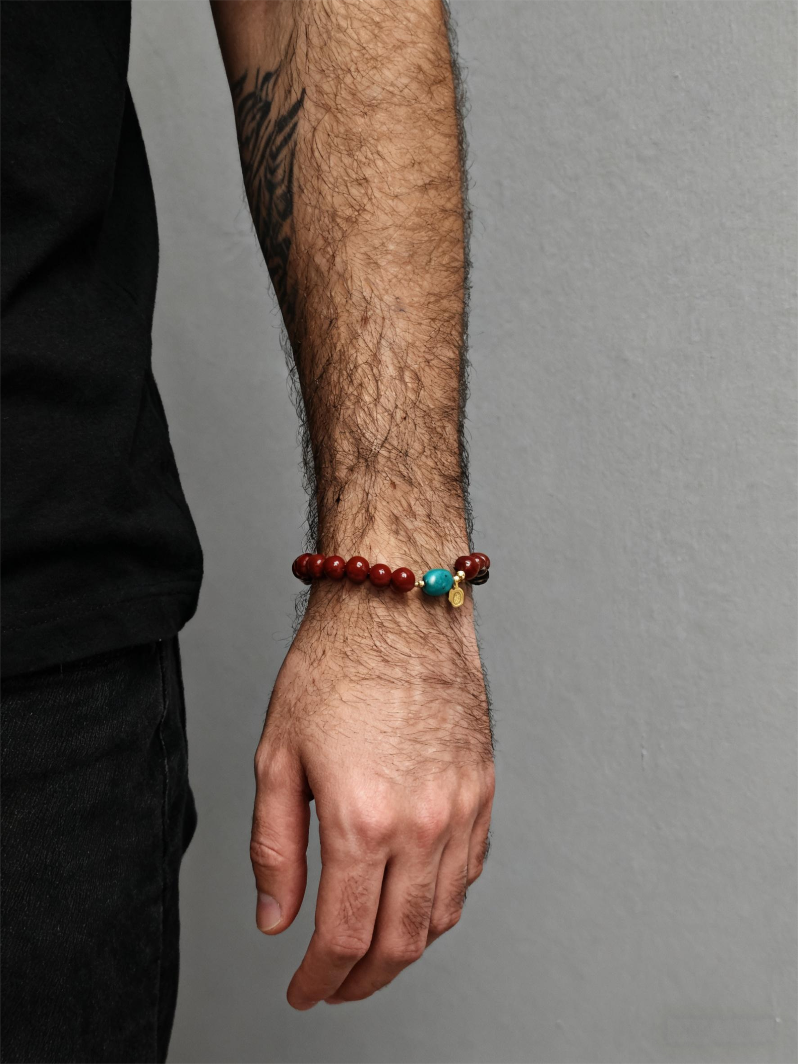 Men’s wrist wearing the Xingtian inspired Steadfast Heart red agate bracelet (10mm) featuring red beads, turquoise centerpiece and gold charm on a grey background