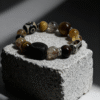 Watchful Relic agate bracelet (12–15mm) with mixed natural stone beads and carved centerpiece, displayed on a textured stone block, luxury handmade myth-inspired jewelry