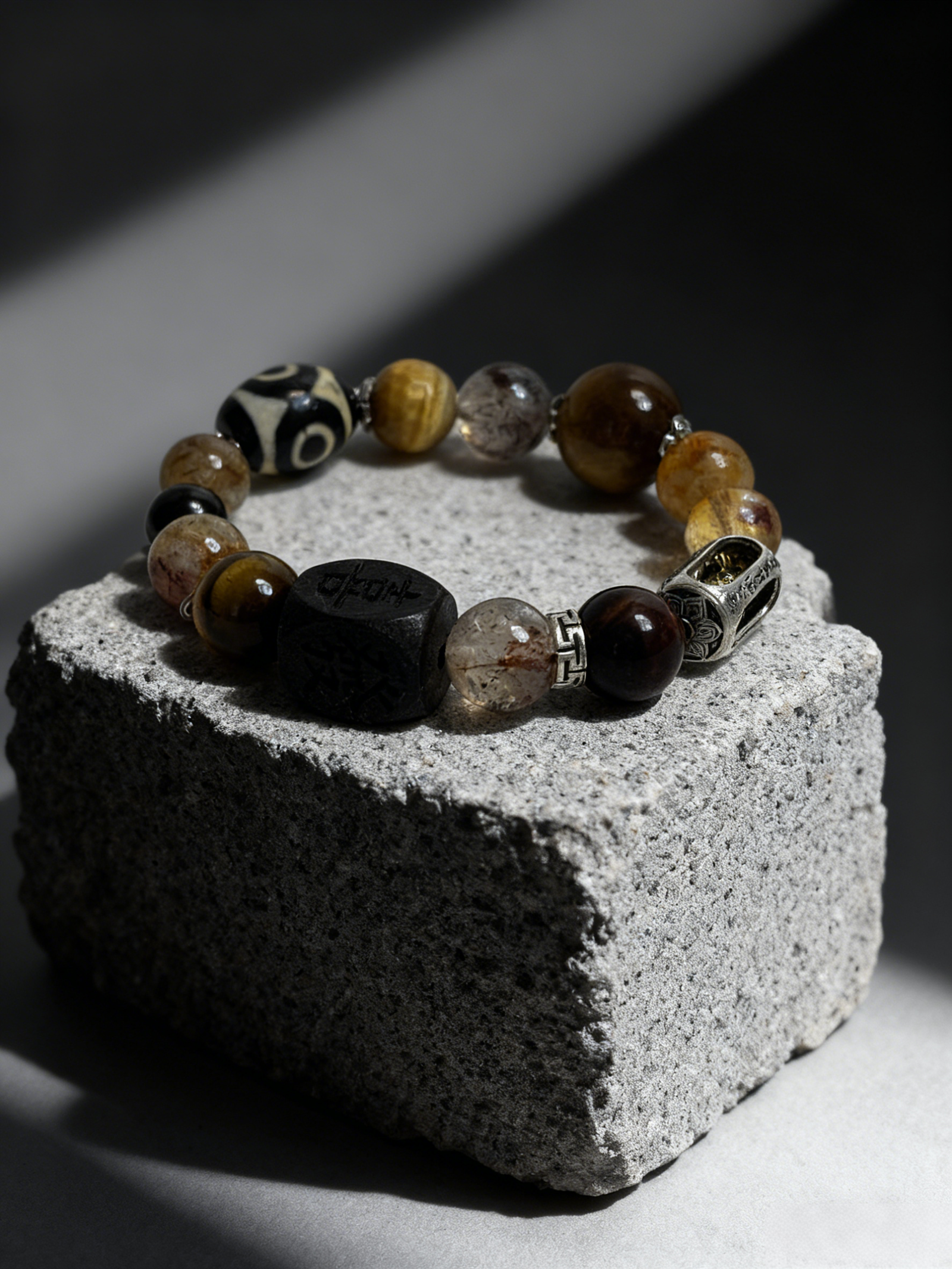Watchful Relic agate bracelet (12–15mm) with mixed natural stone beads and carved centerpiece, displayed on a textured stone block, luxury handmade myth-inspired jewelry