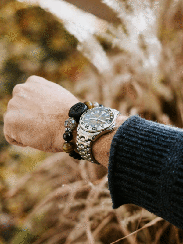 Close-up of the Watchful Relic Agate bracelet worn on a wrist beside a stainless steel watch, featuring mixed natural stone beads and a matte black centerpiece.