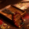 Close-up of Xingtian’s Scarlet Agate Bracelet showing 10mm red agate beads, engraved central emblem, gold charms, and multicolor tassel on a rustic wooden tray