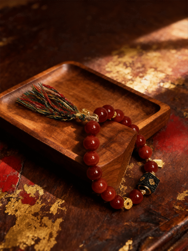 Close-up of Xingtian’s Scarlet Agate Bracelet showing 10mm red agate beads, engraved central emblem, gold charms, and multicolor tassel on a rustic wooden tray