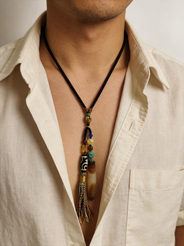 Model wearing the Primordial Layered Agate & Dzi pendant necklace, featuring layered agate, Dzi bead, and multicolor tassel on a black cord, styled with an open white shirt