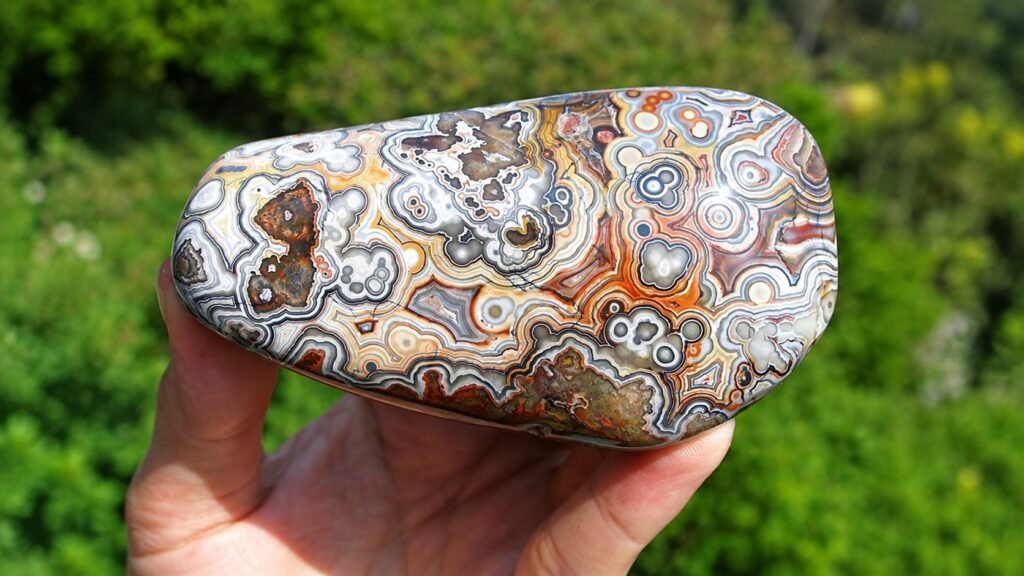Polished Crazy Lace Agate stone with intricate white, gray, orange, and brown banded patterns, held in a hand against a green natural background.